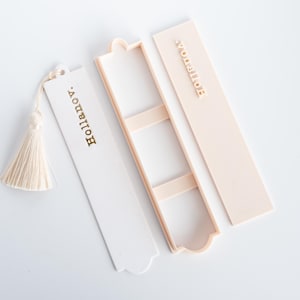 以下が含まれることがあります： 白と淡い桃色の3つのブックマークのセット。1つの白いブックマークにはタッセルと金色の「Hollianov」という文字があります。他の2つのブックマークは淡い桃色で、「Hollianov」という文字が金色で書かれています。