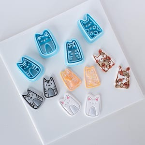 May include: A selection of cat-shaped cookie cutters and cat-themed embellishments. The cutters are blue, and the embellishments feature various colours and patterns, including stripes, spots, and solid colours. The items are arranged on a white surface.