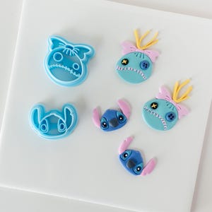 Może przedstawiać: Figurki z lukru w kolorze niebieskim i różowym przedstawiające Stitch'a z filmu Lilo & Stitch. Figurki są ozdobione żółtymi, różowymi i niebieskimi detalami. Są też dwa niebieskie plastikowe wykrawacze do ciasteczek w kształcie głowy Stitch'a.