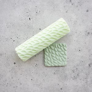 Pode incluir: Um rolo cilíndrico de cor verde-claro com um padrão repetitivo de folhas em relevo, acompanhado por um pequeno azulejo quadrado do mesmo material verde-claro que apresenta uma textura de folha idêntica em toda a sua superfície plana.