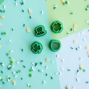 Op de afbeelding: Vier groene plastic koekjesvormpjes in de vorm van een kabouterhoed, een klavertjevier, een klaverblad met drie blaadjes en een pot met goud. De vormpjes liggen in een groepje, omringd door groene, witte en gele strooisels.