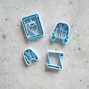 May include: Four blue plastic cookie cutters, shaped like a playing card with a heart, a robe, a scroll, and a robe.