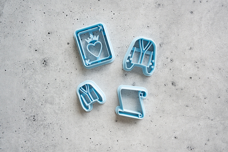 May include: Four blue plastic cookie cutters, shaped like a playing card with a heart, a robe, a scroll, and a robe.