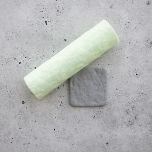 May include: A pale green cylindrical roller with a textured surface positioned diagonally next to a small, square, dark grey tile featuring a matching textured finish. Both items exhibit a matte appearance with subtle, irregular surface patterns.