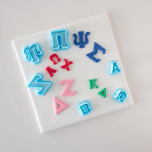 Puede incluir: Una colección de varios cortadores de galletas de plástico con formas de letras y símbolos griegos. El conjunto incluye piezas de color azul claro, azul oscuro, rojo, rosa y verde dispuestas en círculo, con caracteres como Pi, Sigma, Delta y Omega.