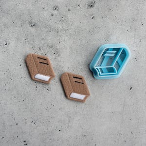 Puede incluir: Dos pequeños recortes en forma de libro de color marrón con bordes de página blancos y detalles de líneas negras, acompañados de un cortador de galletas de plástico azul claro con forma de libro y líneas estructurales internas.
