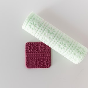 May include: A cylindrical mint green rolling pin with a raised snowflake and geometric pattern, positioned next to a square piece of dark maroon clay that features the same embossed snowflake and geometric design imprinted on its surface.