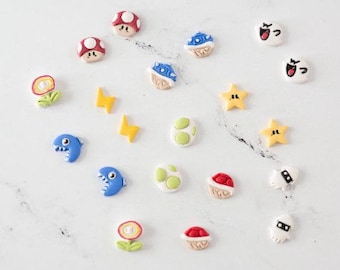 May include: A collection of small, colorful clay charms featuring various video game icons, including red and white mushrooms, blue shells, yellow stars, green-spotted eggs, blue piranha plants, flowers, and white ghost characters arranged on a surface.