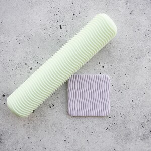 Pode incluir: Um rolo cilíndrico de cor verde-pálido com uma superfície texturizada ondulada ao lado de um pequeno azulejo quadrado de cor roxa clara, apresentando o mesmo padrão ondulado vertical e consistente em toda a sua superfície plana.