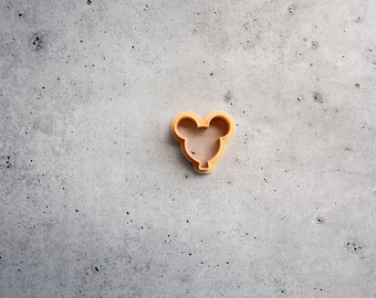 May include: A light orange cookie cutter shaped like a mouse head with two rounded ears and a circular face. The tool features a hollow center and a defined rim for cutting dough into a specific silhouette. The object is solid in color and texture.