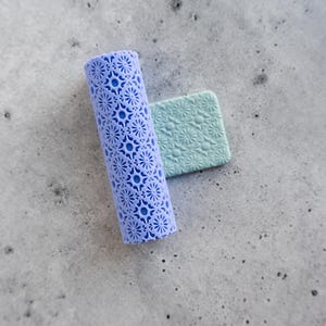 May include: A cylindrical lavender roller tool featuring a detailed starburst and geometric pattern, positioned next to a small, square, light green slab that displays the same embossed floral-inspired design pressed into its surface.