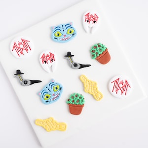 May include: A collection of colorful, whimsical resin embellishments on a white surface. The designs include a blue cat face, a plant in a pot, a yellow shape, and white circles with red text. The embellishments are ideal for crafting.