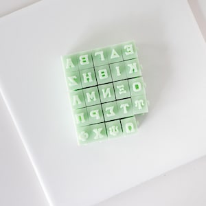 May include: A set of 26 green alphabet letter stamps with white lettering. The stamps are arranged in a grid pattern.