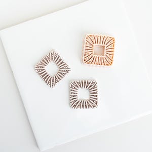 May include: Three square-shaped cookie cutters with intricate, radiating line patterns. One cutter is a light peach color, while the other two are a muted, dusty mauve shade. Each cutter features a hollow center and a detailed, textured design.