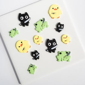 May include: A collection of colorful, cartoonish charms. The charms include black cat-like figures, yellow chicks, and green frogs, each holding a small knife. The charms are arranged on a white surface.
