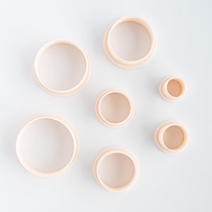 May include: A collection of seven light peach-colored, circular cookie cutters of varying sizes. The cutters are arranged on a white surface, with a raised edge on the bottom. The sizes range from small to large.
