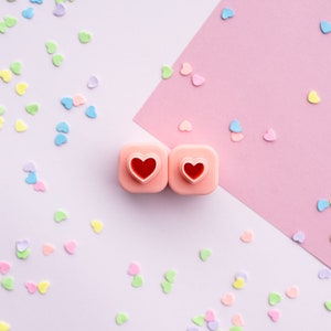 May include: Two pink heart-shaped cookie cutters with red hearts inside, on a pink and white background with pastel pink heart sprinkles.