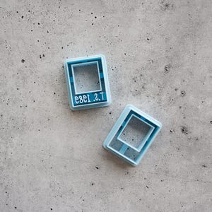 Op de afbeelding: Twee lichtblauwe rechthoekige koekjesvormen. Eén vorm is voorzien van de in reliëf aangebrachte tekst 1989 T.S. in een schreeflettertype. Beide voorwerpen hebben een hol midden en een opstaande buitenrand voor het snijden van vormen.