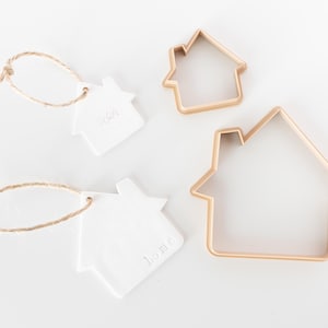 May include: Two white ceramic house-shaped ornaments with jute twine loops, one stamped with 2024 and the other with the word home, alongside two light-colored plastic house-shaped cookie cutters of different sizes on a plain white background.