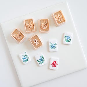 May include: A collection of five light peach-colored plastic cookie cutters featuring floral and character designs, alongside four white rectangular mahjong tiles decorated with blue, green, and red floral and Chinese character illustrations.