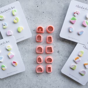 Peut inclure: Quatre ensembles de boucles d'oreilles en argile colorées sur des cartes blanches avec le texte "elizabeth lee clay" et le hashtag "#elizabethleeclay". Un ensemble d'emporte-pièces en argile plastique rose se trouve au centre de l'image.