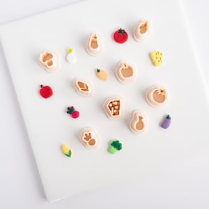 Könnte beinhalten: Eine Sammlung farbenfroher Ausstecher in Lebensmittelform und Miniatur-Lebensmittel. Die Ausstecher sind beige, und die Lebensmittel umfassen eine rote Tomate, einen gelben Käsekeil und eine lila Aubergine. Die Artikel sind auf einer weißen Oberfläche angeordnet.