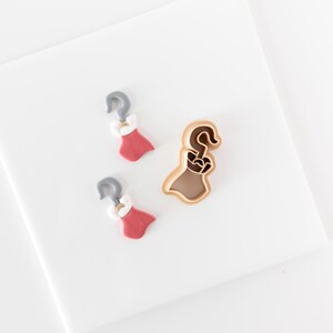 May include: Two small, flat, pink and white dress-shaped charms with gray hook tops, alongside one larger, three-dimensional, light brown plastic cookie cutter in the shape of a dress with a hook top, all arranged on a plain white surface.