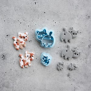 Op de afbeelding: Een verzameling katvormige objecten, waaronder twee kleifiguren met een lapjeskatpatroon, twee lichtblauwe plastic uitsteekvormpjes, twee effen grijze kleifiguren en twee kleine grijze kleibedels, allemaal met een katten-silhouet.