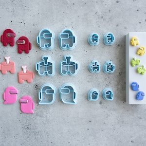 May include: A set of 15 blue and pink plastic cookie cutters shaped like the characters from the video game Among Us. The cutters are arranged in three rows on a gray surface. There is also a white card with five blue and yellow plastic cookie cutters shaped like the characters from the video game Among Us.