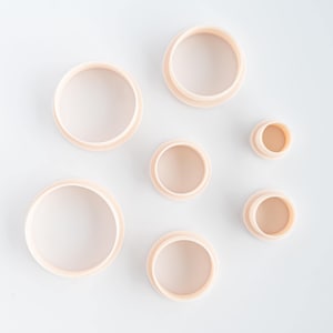 May include: A set of six circular cutters in varying sizes, each featuring a pale peach-colored plastic construction with a raised rim and a hollow center, arranged in a scattered formation on a plain, light-colored surface.