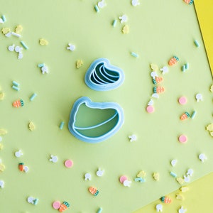 May include: Two blue plastic cookie cutters shaped like an Easter egg and a bunny rabbit on a green background with pastel colored sprinkles.
