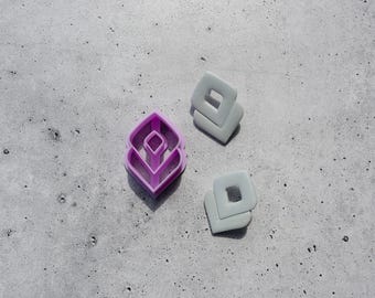 May include: A purple leaf-shaped clay cutter with a central diamond cutout and two light grey square clay pieces, each featuring a centred square hole, arranged on a textured surface. The cutter has a distinct geometric frame design.
