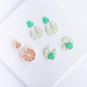 May include: A set of light green frog-shaped clay pieces and pale green circular lily pad shapes with segmented lines, alongside two light pink plastic cutters, one shaped like a small frog and one shaped like a circular citrus slice.