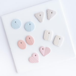 May include: A collection of handmade polymer clay earrings in various shapes: circles, ovals, squares, triangles, and hearts. Each earring is a pastel color, including blue, pink, and beige, with a striped design and a small hole for hanging.