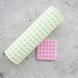 Pode incluir: Um rolo cilíndrico verde-claro com um padrão de textura de pirâmide em relevo, ao lado de uma pequena peça quadrada rosa plana com uma textura de grade piramidal em relevo correspondente, para criar impressões em materiais macios.