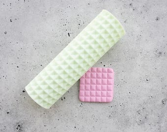 Può includere: Un mattarello cilindrico verde pallido con un motivo a piramide in rilievo, accompagnato da un piccolo pezzo quadrato rosa piatto con una texture a griglia piramidale in rilievo abbinata, per creare impronte su materiali morbidi.