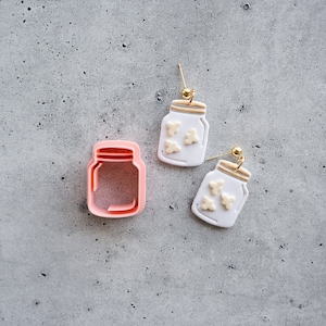 Puede incluir: Un par de pendientes de tarro de albañil blanco con detalles dorados y adornos blancos, junto a un cortador de galletas de tarro de albañil rosa.