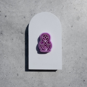 May include: A purple and white nesting doll pin. The doll is outlined in white and has a heart on its chest.