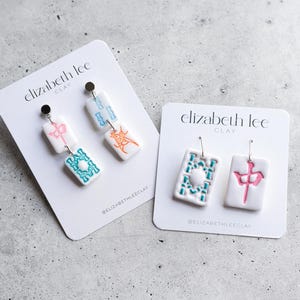 Puede incluir: Pendientes de arcilla blanca con dise&ntilde;os coloridos. Un par presenta un dise&ntilde;o rosa y turquesa, el otro un dise&ntilde;o turquesa y naranja. Los pendientes est&aacute;n en tarjetas blancas con el texto "elizabeth lee CLAY".