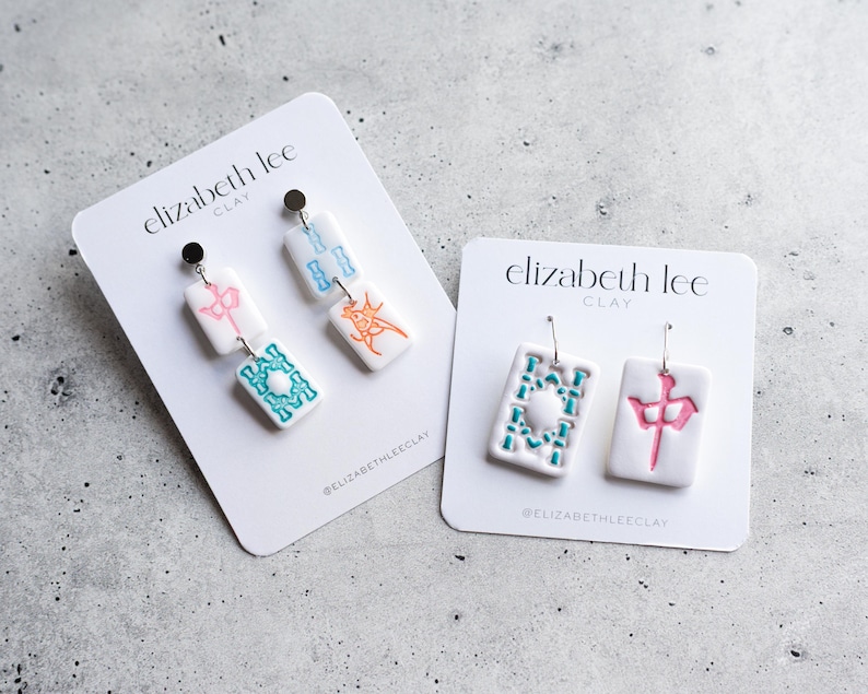 Puede incluir: Pendientes de arcilla blanca con dise&ntilde;os coloridos. Un par presenta un dise&ntilde;o rosa y turquesa, el otro un dise&ntilde;o turquesa y naranja. Los pendientes est&aacute;n en tarjetas blancas con el texto "elizabeth lee CLAY".