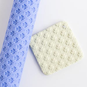 May include: A light blue rolling pin and a square, off-white textured stamp, both featuring a clover pattern. The rolling pin is cylindrical, and the stamp is square. The clover pattern is repeated across both items.