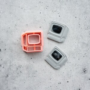 Op de afbeelding: Een perzikkleurige plastic koekjesvorm in de vorm van een vintage computermonitor, vergezeld door twee grijze computer-vormige koekjes met donkere centra, elk met een schermomtrek en een basis die lijkt op een klassieke desktopcomputer.