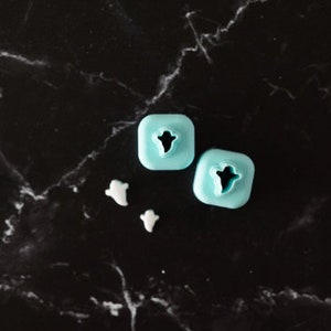 May include: Two light blue plastic ghost-shaped cookie cutters on a black marble surface. Two white ghost-shaped cookies are also on the surface.