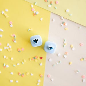 May include: Two light blue square-shaped craft punches featuring small cutouts on their top surfaces, surrounded by a scattered assortment of tiny, colorful, multi-shaped confetti pieces on a split yellow and cream-colored background.