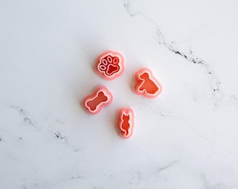 May include: Four pink plastic cookie cutters in the shapes of a paw print, a bone, a sitting dog, and a cat. Each cutter features a raised edge for cutting and a detailed inner impression design for creating shapes in dough or other soft materials.