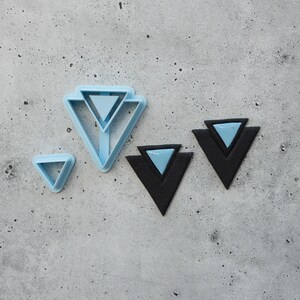 Pode incluir: Dois cortadores triangulares de plástico azul-claro e dois brincos triangulares pretos com incrustações triangulares azul-claro. Os cortadores apresentam um design de moldura geométrica, enquanto os brincos consistem numa base triangular preta grande com um triângulo azul-claro mais pequeno centrado por cima.