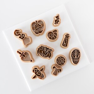 May include: A collection of eleven light brown plastic cookie cutters in various shapes, including a skull-shaped apple, a pirate hook, a seashell, a potion bottle, a dagger, a cat face, a planet, an alien head, a beetle, and a mushroom.