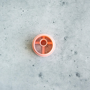 May include: A circular peach-coloured plastic cookie cutter featuring a central ring supported by four perpendicular spokes that extend to the outer rim. The design is simple and functional, with a smooth finish and a consistent light orange hue.
