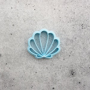 May include: A light blue plastic seashell cookie cutter. The cutter is shaped like a seashell with a scalloped edge.