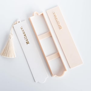 May include: A white rectangular bookmark with a cream-colored tassel and the text "hollanov." in gold, accompanied by a matching cream-colored open storage case and a separate lid featuring the embossed text "hollanov." in a light cream shade.